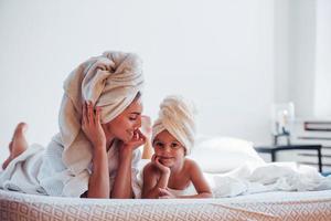 Lying down on white bed together. Young mother with her daugher have beauty day indoors in room photo