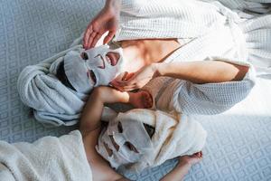 Mother and daughter have spa day with beauty masks on faces. Lying down on bed with towels on heads photo