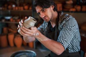 Young ceramist holds fresh handmade pot in hand and looks at results of his work photo