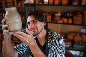 Young ceramist holds fresh handmade pot in hand and looks at results of his work photo