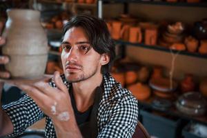 Young ceramist holds fresh handmade pot in hand and looks at results of his work photo