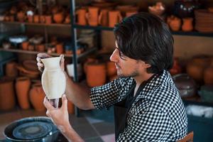 Young ceramist holds fresh handmade pot in hand and looks at results of his work photo