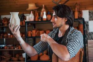 Young ceramist holds fresh handmade pot in hand and looks at results of his work photo