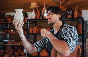 el joven ceramista sostiene una olla fresca hecha a mano en la mano y mira los resultados de su trabajo foto
