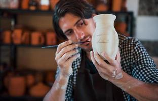 Young ceramist holds his fresh handmade pot and makes some detalizing by special tool stick photo
