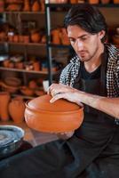 Young ceramist holds fresh handmade product made of clay in hands and looks at results of his work photo