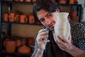 Young ceramist holds his fresh handmade pot and makes some detalizing by special tool stick photo