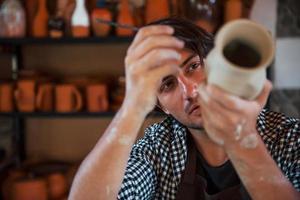 Young ceramist holds his fresh handmade pot and makes some detalizing by special tool stick photo