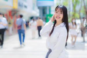 Cute young Asian school girl l who wears white shirt standing and smiling while outdoors in the city with people and office building in the background. photo