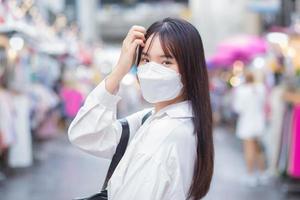 Linda joven estudiante asiática que usa camisa blanca y mascarilla para prevenir enfermedades de pie y sonriendo mientras está al aire libre en la ciudad con la calle con tiendas en el fondo.