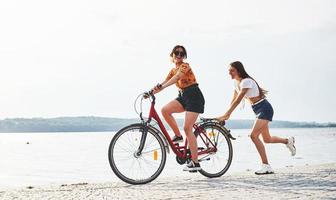 Girl runs near bicycle. Two female friends on the bike have fun at beach near the lake photo