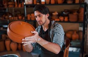 Young ceramist holds fresh handmade product made of clay in hands and looks at results of his work photo