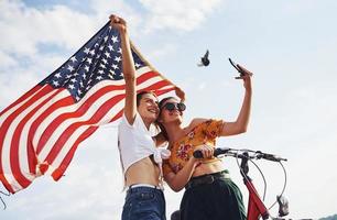 Against cloudy sky. Two patriotic cheerful woman with bike and USA flag in hands makes selfie photo