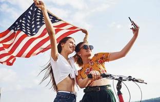 Against cloudy sky. Two patriotic cheerful woman with bike and USA flag in hands makes selfie photo