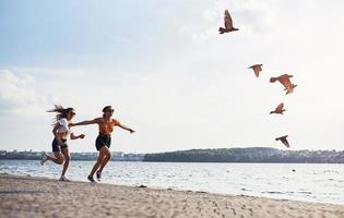 Birds flying up in the air. Two female friends runs and have fun at beach near the lake at sunny daytime photo