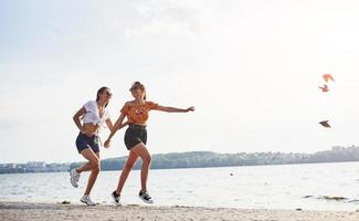 Birds flying up in the air. Two female friends runs and have fun at beach near the lake at sunny daytime photo