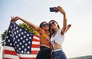 Green tree at background. Two patriotic cheerful women with USA flag in hands making selfie outdoors in park photo