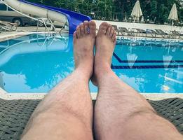 vacaciones en el mar en la playa. turista tomando el sol en una tumbona junto a la piscina. piernas masculinas blancas. piscina con agua turquesa y clorada con un patrón en el fondo foto
