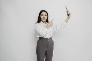 Shocked Asian woman wearing white shirt and holding her phone, isolated by white background photo