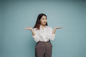 Shocked Asian woman wearing white shirt pointing at the copy space beside her, isolated by blue background photo