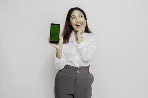 mujer asiática emocionada con pantalones blanco apuntando al espacio de copia a su lado mientras sostiene su teléfono, aislada por fondo blanco foto