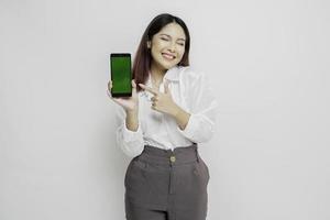 Excited Asian woman wearing white shirt pointing at the copy space beside her while holding her phone, isolated by white background photo