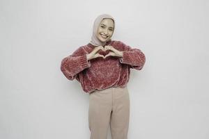 A happy young Asian woman wearing a pink sweater and hijab feels romantic shapes heart gesture expresses tender feelings photo