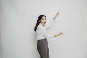 Shocked Asian woman wearing white shirt pointing at the copy space beside her, isolated by white background photo