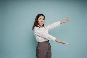 Shocked Asian woman wearing white shirt pointing at the copy space beside her, isolated by blue background photo