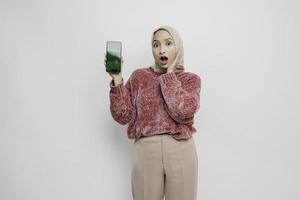 Surprised Asian Muslim woman wearing pink sweater and hijab showing copy space on her smartphone, isolated by white background photo