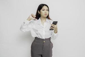 A disappointed Asian woman wearing white shirt gives thumbs down hand gesture of disapproval, isolated by white background photo