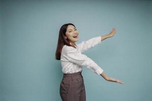 Excited Asian woman wearing white shirt pointing at the copy space beside her, isolated by blue background photo