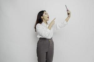 mujer asiática sorprendida con camisa blanca y sosteniendo su teléfono, aislada de fondo blanco foto