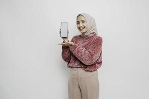 Excited Asian Muslim woman wearing pink sweater and hijab pointing at the copy space beside her while holding her phone, isolated by white background photo