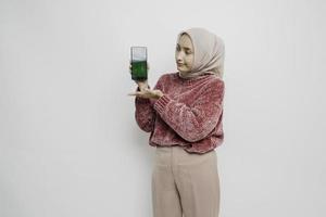 Excited Asian Muslim woman wearing pink sweater and hijab pointing at the copy space beside her while holding her phone, isolated by white background photo