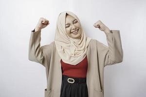 Excited Asian Muslim woman wearing a hijab showing strong gesture by lifting her arms and muscles smiling proudly photo