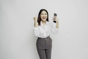 A young Asian woman with a happy successful expression wearing white shirt and holding smartphone isolated by white background photo