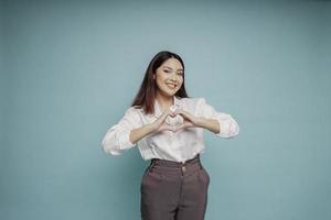 A happy young Asian woman wearing a white shirt feels romantic shapes heart gesture expresses tender feelings photo