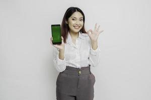 A portrait of a excited Asian woman wearing a white shirt while holding her phone and gesturing approval sign, isolated by white background photo