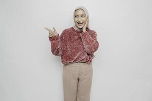 mujer musulmana asiática emocionada con suéter rosa y hiyab señalando el espacio de la copia a su lado, aislada de fondo blanco foto