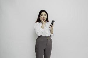 Shocked Asian woman wearing white shirt and holding her phone, isolated by white background photo