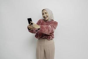 A portrait of a happy Asian woman wearing a pink sweater and hijab, holding her phone, isolated by white background photo