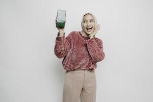 Excited Asian Muslim woman wearing pink sweater and hijab pointing at the copy space beside her while holding her phone, isolated by white background photo