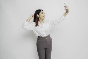 una joven asiática con una expresión feliz y exitosa que usa pantalones blanco y sostiene un teléfono inteligente aislado de fondo blanco foto