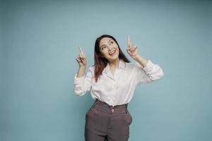 Excited Asian woman wearing white shirt pointing at the copy space on top of her, isolated by blue background photo