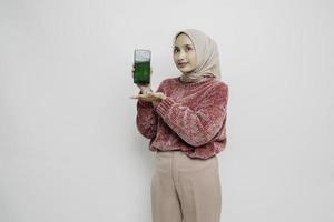 Excited Asian Muslim woman wearing pink sweater and hijab pointing at the copy space beside her while holding her phone, isolated by white background photo