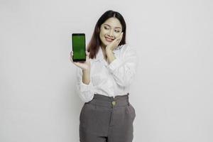 mujer asiática emocionada con pantalones blanco apuntando al espacio de copia a su lado mientras sostiene su teléfono, aislada por fondo blanco foto
