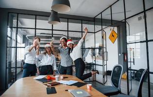 With hands up. Employees in formal clothes, glasses with champagne and in christmas hats celebrating new year in the office photo