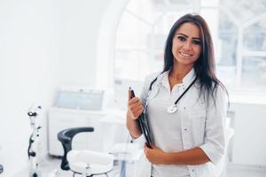 With notepad in hands. Brunette female doctor stands in modern office and looks at camera photo