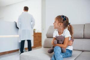 Doctors behind. Cute little girl with teddy bear in hands sits in waiting room of hospital photo